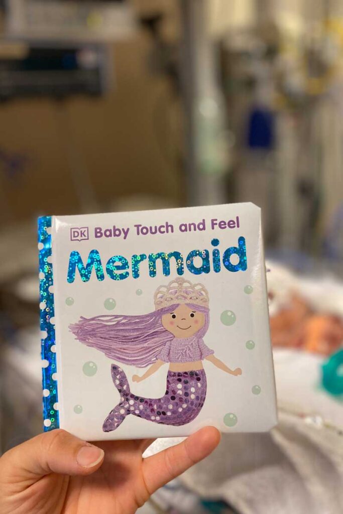 A hand holds a children's Baby Touch and Feel board book titled "Mermaid" in blue shiny letters with bubbles. The cover features a cute illustration of a mermaid with purple hair and a sparkly tail. The blurred background is of a baby's ICU room.