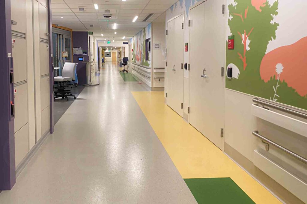 An empty hallway at Boston Children's Hospital. The corridor is brightly lit and features colorful, playful wall art with trees and flowers, creating a welcoming atmosphere. There are chairs and workstations along the sides, with various doors and equipment storage units visible.