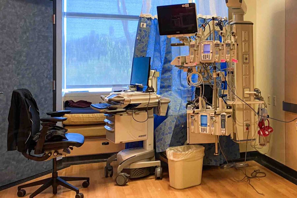 The image shows an empty ICU room in a children's hospital. There is a workstation with a computer on a mobile cart, an office chair, and a complex setup of medical equipment and monitors attached to a wall unit. The room has a blue privacy curtain, a window, and a small built in bench area.