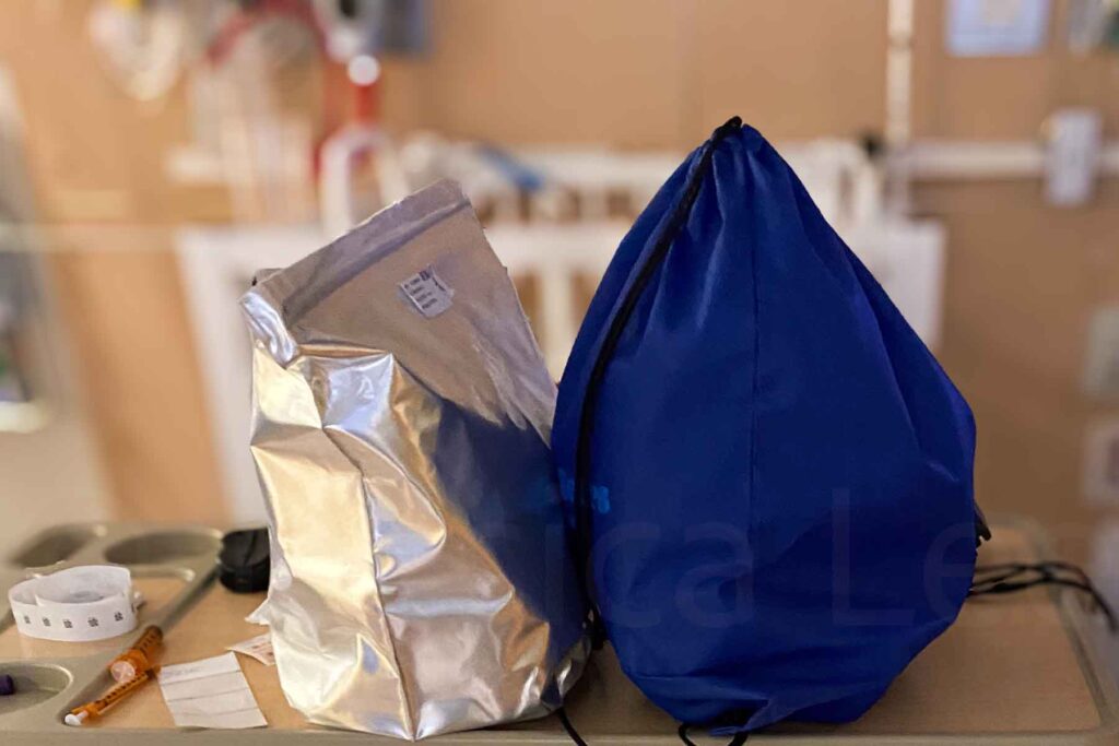 This image shows a silver insulated cooler bag and a large blue drawstring bag placed on a small counter-height table in a hospital room. Also on the table, there is a medical wristband and a syringe.