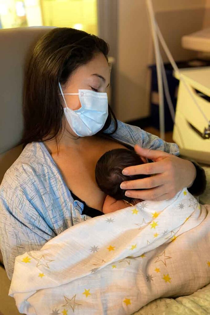 A mother wearing a hospital gown and a blue medical face mask cradles her newborn baby chest to chest wrapped in a white blanket with yellow and gray stars. She is seated in a hospital room with her eyes shut, gently touching the baby's head as the infant sleeps.